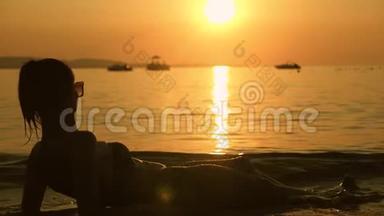 日落时分，美丽的年轻女孩躺在海滩上。 度假的女孩在海滩上放松，看日落。 夏季